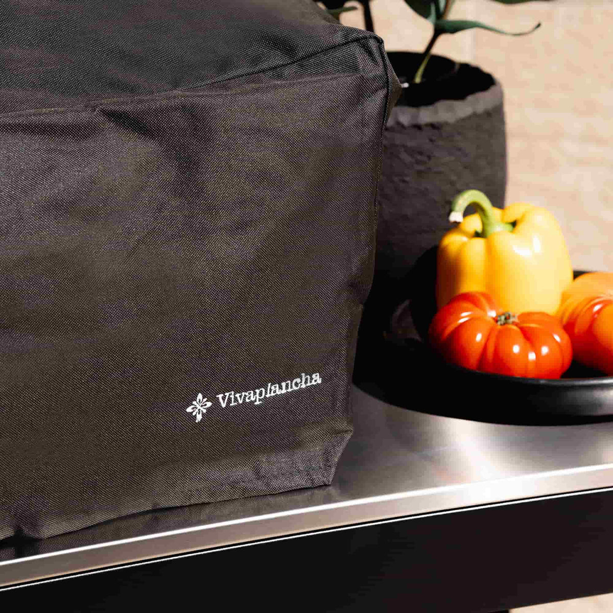 Gros plan sur la housse noire Vivaplancha couvrant une plancha, avec logo visible au premier plan, posée sur une desserte inox et entourée de légumes.