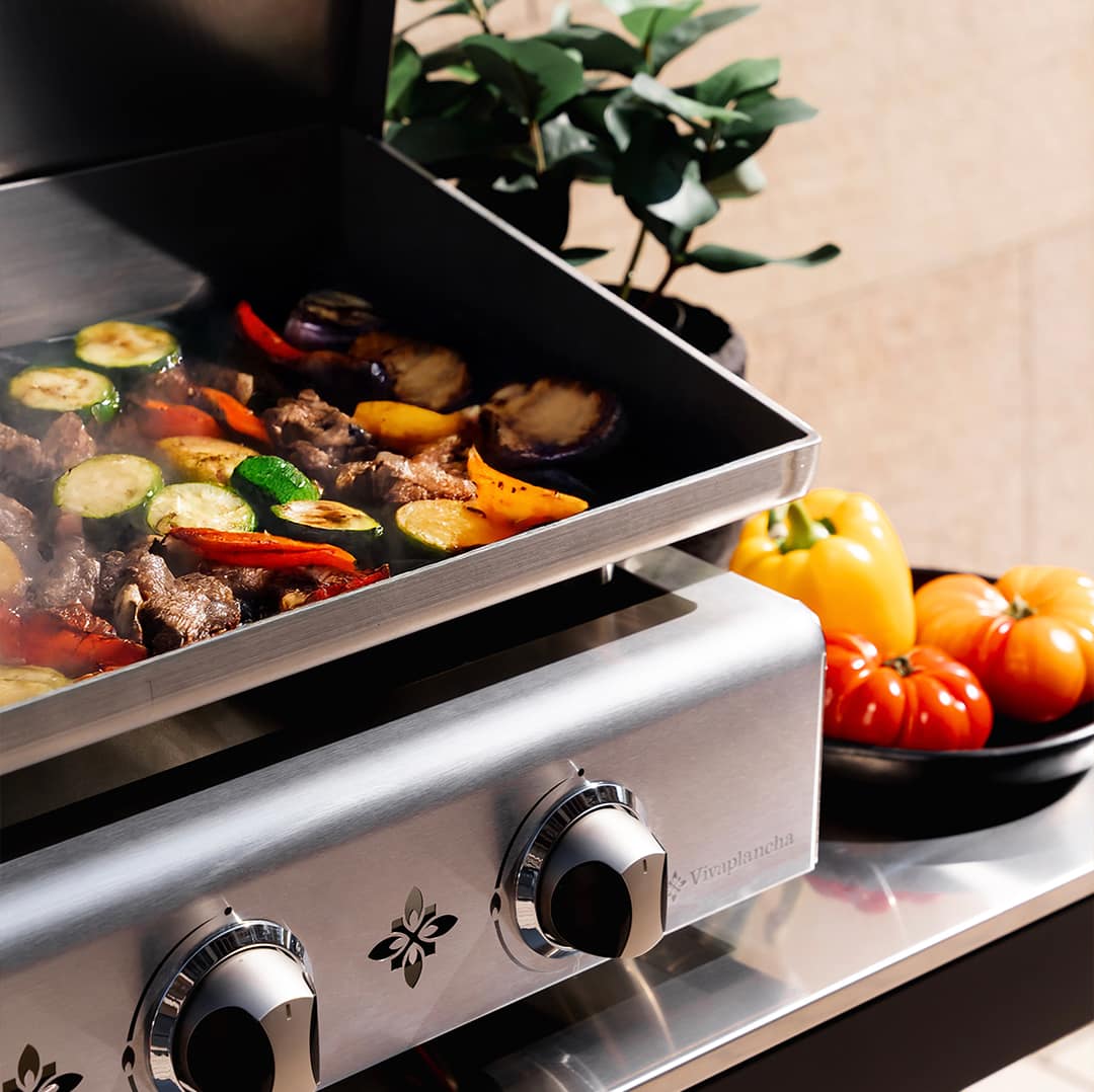 Légumes et morceaux de viande en train de griller sur une plancha inox Vivaplancha, avec de la vapeur et des légumes frais posés à côté.